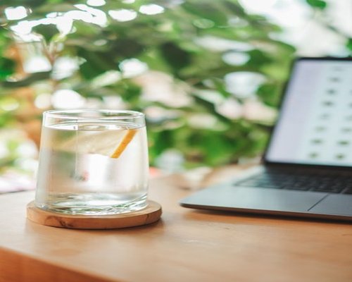 A clear glass of water sitting next to a laptop on a wooden desk bathed in natural light